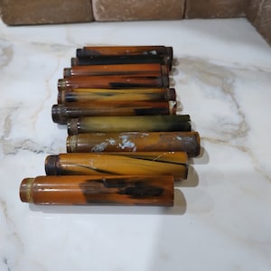 Puede incluir: Una colección de nueve objetos cilíndricos en forma de tubo dispuestos en una fila escalonada. Cada pieza presenta un patrón jaspeado similar al carey en tonos ámbar, marrón, amarillo y negro, con texturas y desgaste superficial variado.