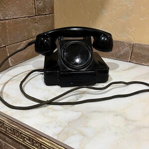 Puede incluir: Un teléfono de disco negro con un cable en espiral. El teléfono está sobre una superficie de mármol blanco con un borde dorado.