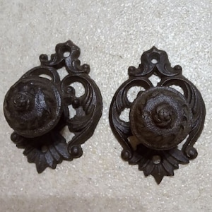 May include: Two black cast iron door knobs with ornate floral designs. The knobs have a raised, circular design in the center.