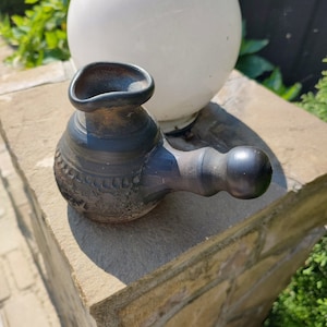 May include: A dark brown ceramic pitcher with a spout and a handle. The pitcher has a textured surface and is decorated with a pattern of small circles.
