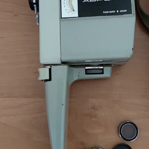 May include: A vintage silver and black 8mm film camera with a black strap. The camera has a dial with numbers 0-7.5 and the text "АВРОРА СДЕЛАНО В СССР" on the front. The camera is on a wooden surface with three round filters: one brown, one black, and one yellow.