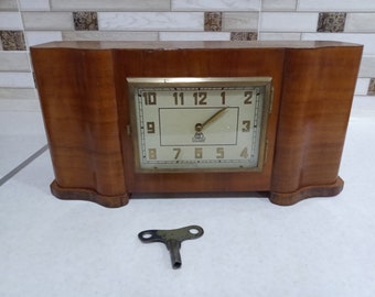 May include: A vintage wooden mantel clock with a white face and black Roman numeral hour markers. The clock has a key wind mechanism and a brass coloured trim. The clock is sitting on a white surface with a key for winding the clock.