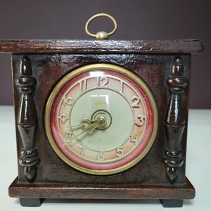 May include: A vintage wooden clock with a red and white face. The clock has a gold-colored trim and a small brass handle on the top. The clock face reads "a.mas".