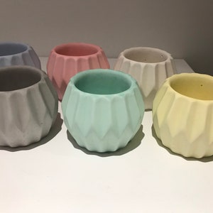 Puede incluir: Seis pequeñas macetas de hormigón geométricas en varios colores pastel, incluyendo azul claro, rosa, gris, verde claro y amarillo. Las macetas tienen un diseño único y facetado.