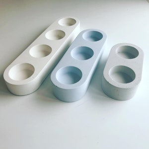 Puede incluir: Tres portavelas ovalados de hormigón. Los portavelas son de color blanco, azul claro y gris. El portavelas blanco tiene cuatro pozos para velas, mientras que los portavelas azul claro y gris tienen tres pozos para velas cada uno.