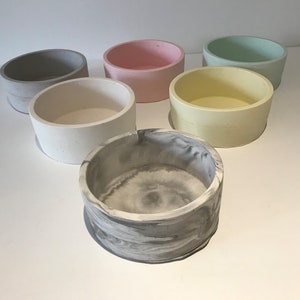 Puede incluir: Seis cuencos de hormigón en varios colores, incluyendo gris, rosa, verde claro, amarillo, blanco y gris marmoleado. Los cuencos son todos del mismo tamaño y forma, y están dispuestos en semicírculo sobre una superficie blanca.