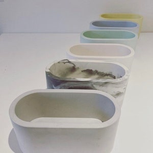 May include: A set of seven oval-shaped concrete planters in various colours, including white, grey, beige, green, blue, and yellow. The planters are arranged in a row from largest to smallest.