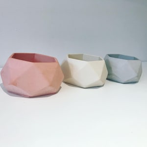 May include: Three geometric concrete planters in pastel shades: pink, off-white, and grey. Each planter features a faceted design and an open top. The planters are arranged in a row on a white surface, ideal for small plants or succulents.
