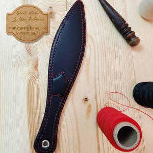 May include: Black leather knife sheath with red stitching. The sheath is on a wooden surface with spools of red and black thread.