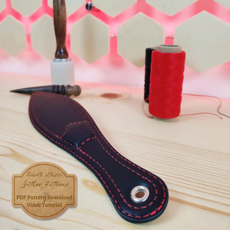 May include: A black leather paddle with red stitching and a metal ring. The paddle is on a wooden surface with two spools of red and black thread.