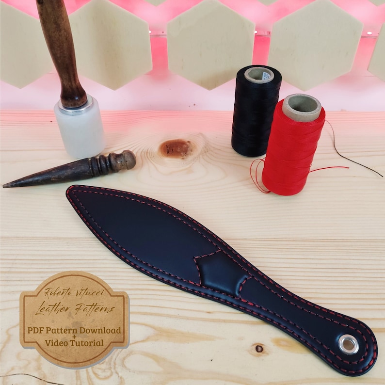 May include: Black leather paddle with red stitching, a wooden mallet, an awl, and two spools of thread, one red and one black. The paddle has a hole at the end and a label that says "Roberto Vitucci Leather Patterns" and "PDF Pattern Download Video Tutorial".