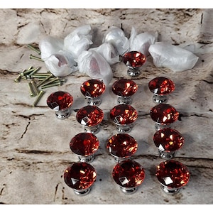 May include: A set of twelve round, faceted red glass cabinet knobs with silver-toned metal bases, arranged in a grid. Several additional knobs are wrapped in white protective plastic, and a collection of silver-toned metal mounting screws is included.