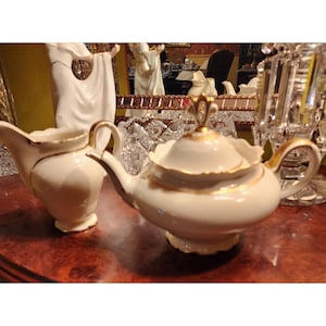 May include: A cream-colored porcelain teapot with a matching lid and a cream pitcher, both featuring delicate gold trim along the edges. The teapot has a scalloped lid with a gold finial, a curved spout, and a handle, while the pitcher has a pouring spout.