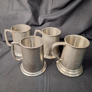 May include: Four vintage pewter tankards with handles. The tankards have a cylindrical shape with a slightly flared base. The metal has a dull, aged appearance. One tankard has an inscription. The tankards are arranged on a dark surface.