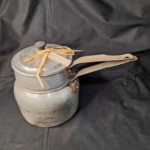 May include: A vintage aluminum double boiler with a lid and long handle. The pot has a worn, aged appearance, with a small tag tied to the lid with raffia. The handle is attached with rivets.
