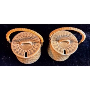 May include: Two matching beige ceramic containers with lids and handles. The lids feature a sunburst design, and the containers have a woven basket-like texture. The handles are a light orange color. The containers are on a black surface.