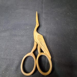 May include: A pair of decorative scissors in the shape of a bird, likely a crane or heron. The scissors are made of a light brown metal, with a detailed design of the bird's feathers and body. The scissor handles are circular.