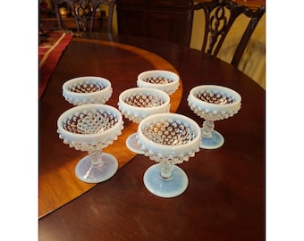 May include: A set of five opalescent glass sherbet dishes featuring a raised hobnail texture on the exterior of the bowls and stems. Each dish has a circular base and a wide, shallow bowl with a scalloped rim, displaying a milky white and blue hue.