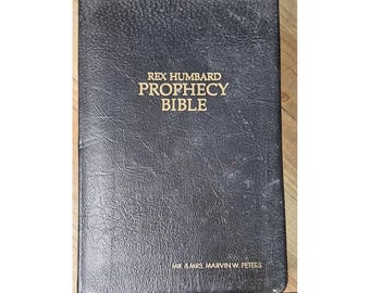 May include: A black, textured, hardcover book titled Rex Humbard Prophecy Bible in gold lettering on the front cover. The bottom of the cover features the gold printed text Mr. & Mrs. Marvin W. Peters. The book has a rectangular shape.