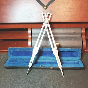 May include: A silver-toned metal drafting compass with two pointed legs and a central adjustment screw, featuring engraved markings along the arms. The tool is positioned in front of an open, rectangular storage case with a blue velvet interior.