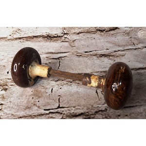 May include: A pair of round, dark brown, glossy ceramic door knobs connected by a central metal spindle. The knobs feature a smooth, rounded surface with visible wear on the off-white, cylindrical neck sections connecting to the metal rod.