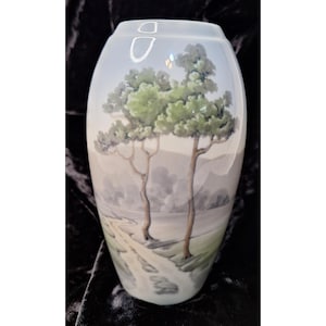 May include: A light blue, oval-shaped vase with a landscape painting. The artwork depicts two trees with green foliage and brown trunks, a winding path, and a distant mountain range. The vase is a decorative piece.