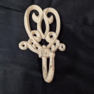May include: Off-white, ornate metal wall hook with a decorative heart-shaped top and a curved hook. The hook has a distressed, vintage appearance, with visible wear and tear. The design includes scrollwork and a central oval opening.