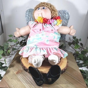 May include: A doll with light skin, blue eyes, and brown yarn hair wearing a pink dress with a light blue floral pattern, a colorful flower lei, white lace-trimmed socks, and white fabric shoes, accompanied by a pair of black shoes.