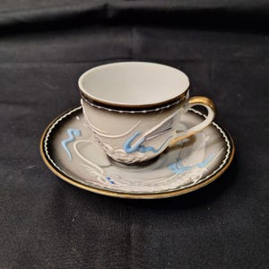 May include: A vintage teacup and saucer set. The cup and saucer are gray with a black border and gold trim. The set features white and blue dragon designs. The cup handle is gold.
