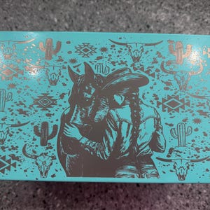May include: A turquoise box with a western theme. The box features a brown illustration of a person in a cowboy hat embracing a horse. The background is decorated with cacti, bull skulls, and geometric patterns in a metallic gold color.
