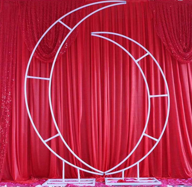 May include: White metal archway shaped like two half moons, standing in front of a red fabric backdrop.