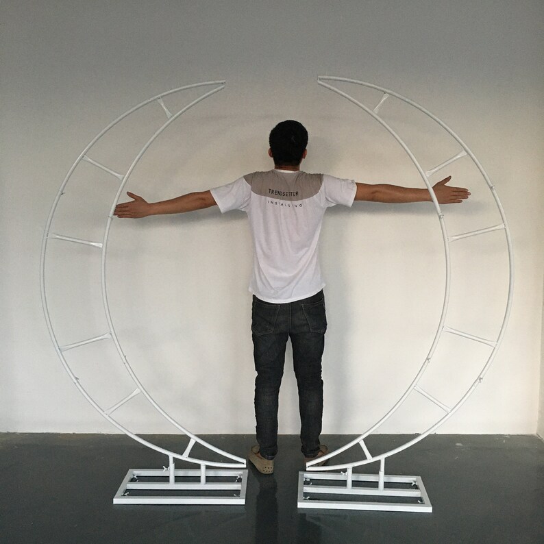 May include: White metal archway shaped like a crescent moon with a person standing in front of it. The archway has a series of horizontal bars across the curved section.