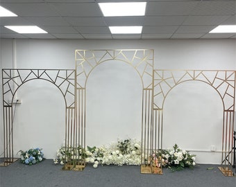 May include: Three gold metal archways with geometric designs. The arches are freestanding and have a modern style. They are decorated with white and pink flowers.