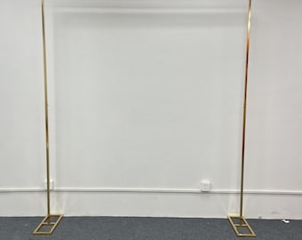 May include: A gold metal archway stand with a rectangular base. The stand is freestanding and is made of thin metal tubing.