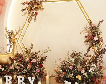 May include: A gold hexagonal archway decorated with floral arrangements, surrounded by greenery and white letters spelling "marry me".
