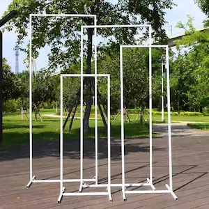 Op de afbeelding: Verschillende witte rechthoekige metalen frames van verschillende afmetingen zijn buiten geplaatst. De frames staan op een houten terras met een achtergrond van bomen. De frames zijn ontworpen om rechtop te staan en worden ondersteund door een basis.