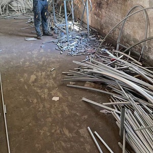 May include: A man wearing a blue shirt and jeans uses a grinder to cut metal pipes in a workshop. Sparks fly from the grinder as he works. The floor is covered in metal scraps and pipes.