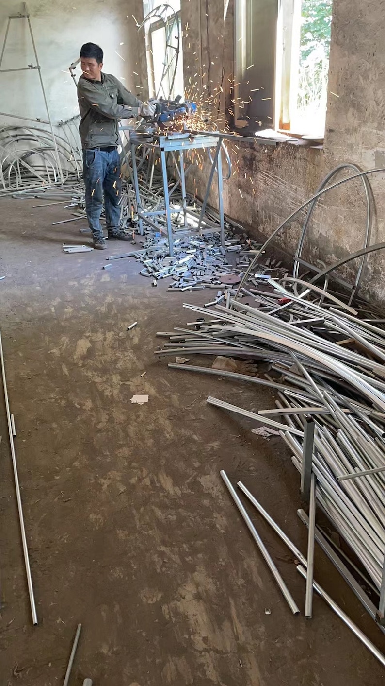 May include: A man wearing a blue shirt and jeans uses a grinder to cut metal pipes in a workshop. Sparks fly from the grinder as he works. The floor is covered in metal scraps and pipes.