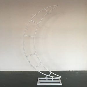 May include: White metal arch shaped like a crescent moon. The arch has a square base and is designed for use as a wedding or event backdrop.