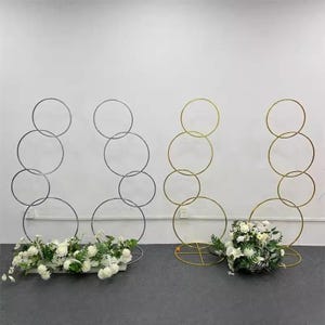 May include: Two metal circle arch backdrops, one silver and one gold, are displayed. Each arch is adorned with white flowers. The arches are designed for events and weddings, providing a decorative backdrop for photos or ceremonies.