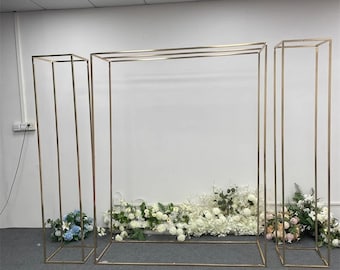 May include: A gold metal archway backdrop with two matching tall rectangular frames. The archway is decorated with white and pink flowers.