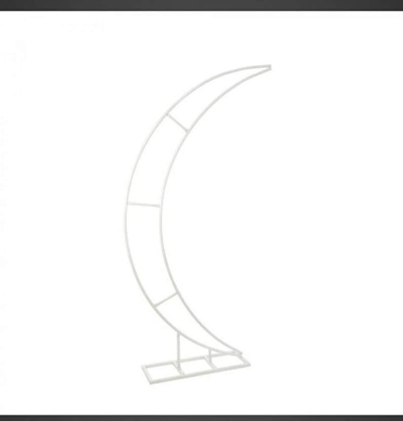 May include: White metal crescent moon arch stand for wedding or event decor. The stand is made of metal tubing and has a simple, modern design.
