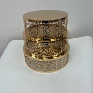 May include: A three-tiered, gold-colored decorative storage set. Each tier is a different size, with a solid top and a patterned, openwork design on the sides. The set is made of metal and has a reflective surface.