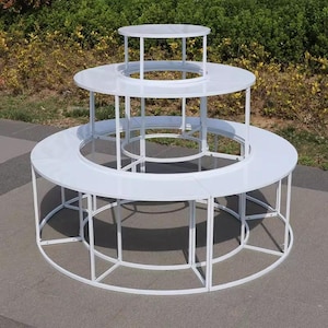 May include: A tiered, white display stand with three circular platforms of varying sizes. The stand has a minimalist design with a white metal frame and is suitable for showcasing products. The platforms are connected by vertical supports.