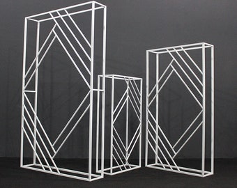 May include: Three white metal geometric arch backdrops with a diamond pattern. The backdrops are different sizes and can be used for weddings, parties, or other events.