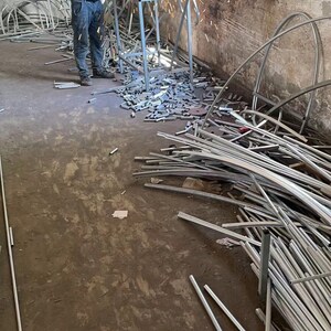 May include: A man wearing a blue shirt and jeans is using a grinder to cut metal pipes. Sparks are flying from the grinder. There are many metal pipes scattered on the floor.