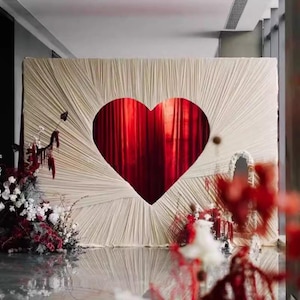 Könnte beinhalten: Ein cremefarbener Hintergrund mit einem großen herzförmigen Ausschnitt, der einen roten Vorhang freigibt. Der Hintergrund hat ein strukturiertes, radiales Design. Blumengestecke mit roten und weißen Blumen befinden sich auf der linken und rechten Seite und schaffen eine romantische Atmosphäre.