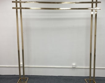 May include: Gold metal archway with two horizontal bars. The archway is freestanding and has a square base.