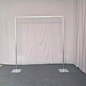 以下が含まれることがあります： 結婚式やイベントに最適な、シルバーの金属製スクエアアーチ。アーチは長方形の上面と2つの垂直サポートがあり、それぞれ正方形のベースで固定されています。白い背景に対して、灰色のカーペットの上に立っています。