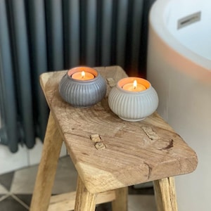 May include: Two small ceramic candle holders with ribbed detailing, one grey and one white, with lit tea lights inside, sitting on a rustic wooden stool.